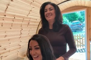 Karis Petersen standing behind a seated woman with freshly styled hair inside the wooden Karistyle salon pod at Otham